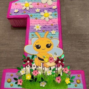 Pu&ograve; includere: Una pi&ntilde;ata a forma di numero uno rosa e viola con un'ape e dei fiori. La pi&ntilde;ata &egrave; decorata con glitter e ha una corda attaccata alla parte superiore.