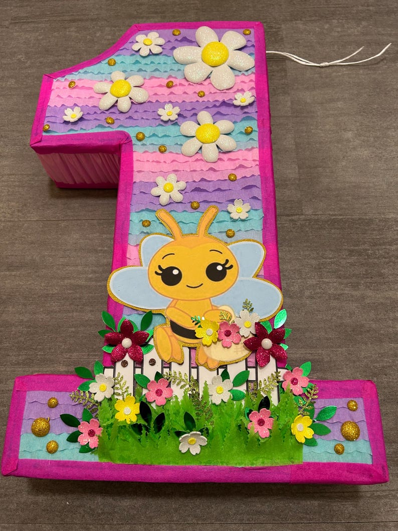 Pu&ograve; includere: Una pi&ntilde;ata a forma di numero uno rosa e viola con un'ape e dei fiori. La pi&ntilde;ata &egrave; decorata con glitter e ha una corda attaccata alla parte superiore.
