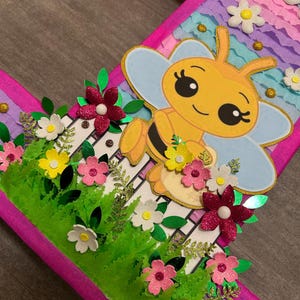 Pu&ograve; includere: Un numero uno di cartone rosa e viola con un'ape e dei fiori. L'ape &egrave; gialla con strisce nere e ha ali blu e bianche. I fiori sono rosa, bianchi e gialli. Il numero uno &egrave; decorato con foglie verdi e glitter.