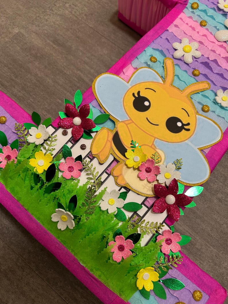 Pu&ograve; includere: Un numero uno di cartone rosa e viola con un'ape e dei fiori. L'ape &egrave; gialla con strisce nere e ha ali blu e bianche. I fiori sono rosa, bianchi e gialli. Il numero uno &egrave; decorato con foglie verdi e glitter.