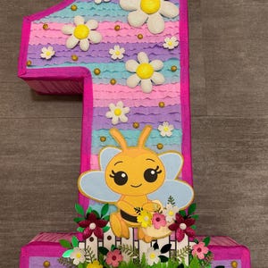 Pu&ograve; includere: Una pi&ntilde;ata a forma di numero uno rosa e blu con un designo di ape. La pi&ntilde;ata &egrave; decorata con margherite bianche, glitter dorato e foglie verdi. L'ape tiene un cestino di fiori.