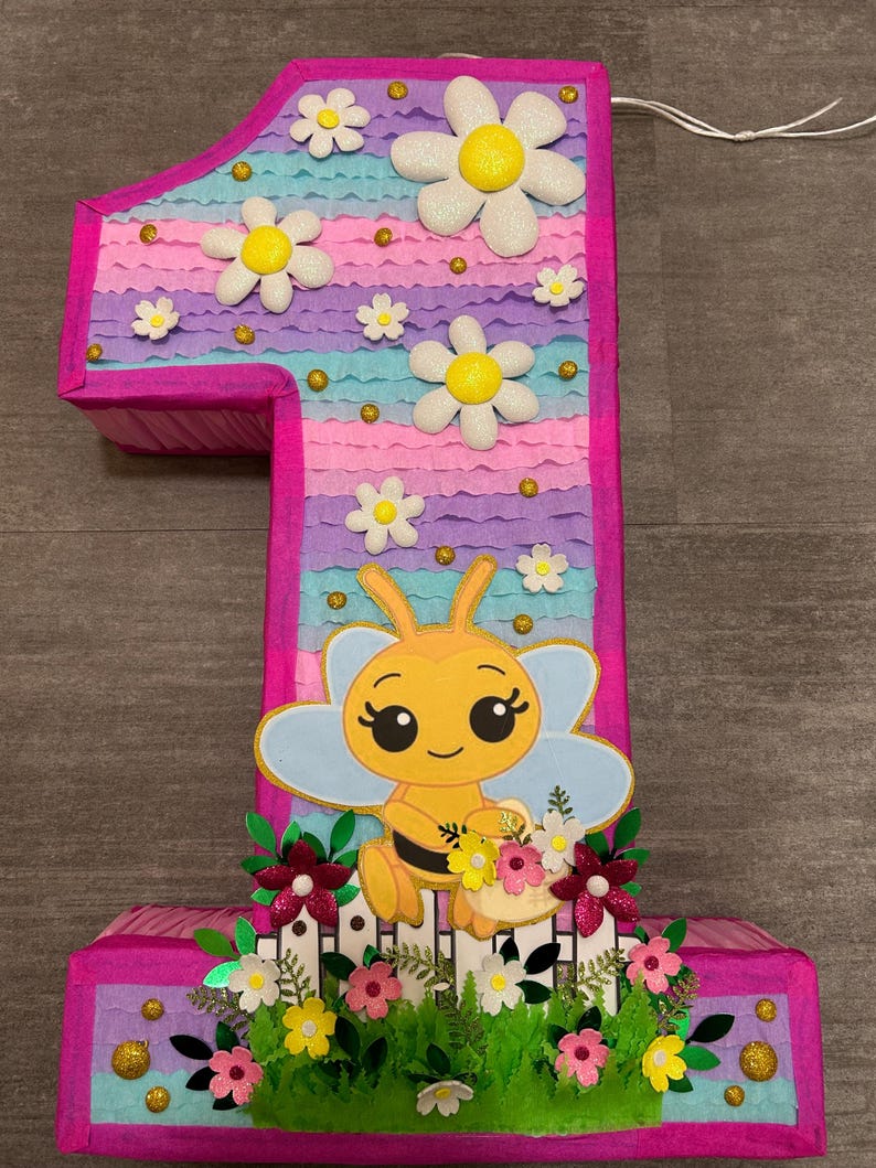 Pu&ograve; includere: Una pi&ntilde;ata a forma di numero uno rosa e blu con un designo di ape. La pi&ntilde;ata &egrave; decorata con margherite bianche, glitter dorato e foglie verdi. L'ape tiene un cestino di fiori.
