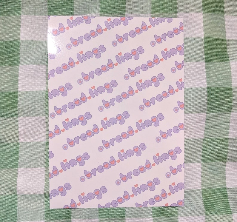 K&ouml;nnte beinhalten: Ein wei&szlig;er Hintergrund mit einem sich wiederholenden Muster aus violettem Text, der "bread.lings" sagt, mit einem rosa Herzen in der Mitte des Wortes "bread".