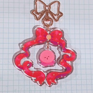 May include: A clear acrylic keyring with a pink wreath design, red ribbon, and a smiling pink character. It has a rose gold bow and a small charm. The background is a grid pattern.