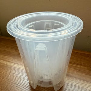 May include: A clear plastic plant pot with a wide, tiered rim. The pot has a conical shape with vertical supports and a series of drainage holes at the bottom. The pot is designed for indoor or outdoor use.