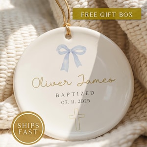 May include: A white ceramic baptism ornament with a light blue bow illustration, the name "Oliver James", the word "BAPTIZED", and the date "07. 11. 2025" in gold lettering. A gold cross is at the bottom. A gold cord is attached for hanging.