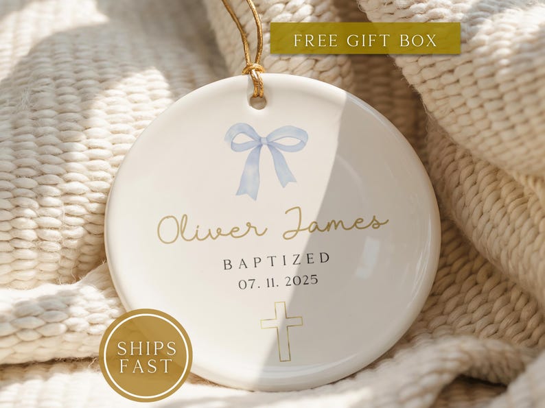 May include: A white ceramic baptism ornament with a light blue bow illustration, the name "Oliver James", the word "BAPTIZED", and the date "07. 11. 2025" in gold lettering. A gold cross is at the bottom. A gold cord is attached for hanging.