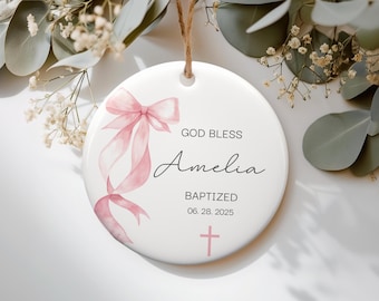 May include: A white, round ceramic ornament with a pink ribbon design and the text "GOD BLESS Amelia BAPTIZED 06.28.2025" in black. A pink cross is at the bottom. The ornament hangs from a natural twine string.