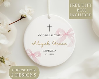 May include: A white ceramic baptism ornament with a cross and the text "GOD BLESS YOU" and "Aliyah Grace". It features pink watercolor bows and the date 07.11.2025. The ornament includes the text "CHOOSE FROM 2 DESIGNS" and "FREE GIFT BOX INCLUDED".