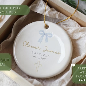 May include: A white ceramic ornament with a gold string, featuring the name "Oliver James" and the date "07. 11. 2025". It includes a light blue bow and a cross design. The ornament is packaged in a gift box.
