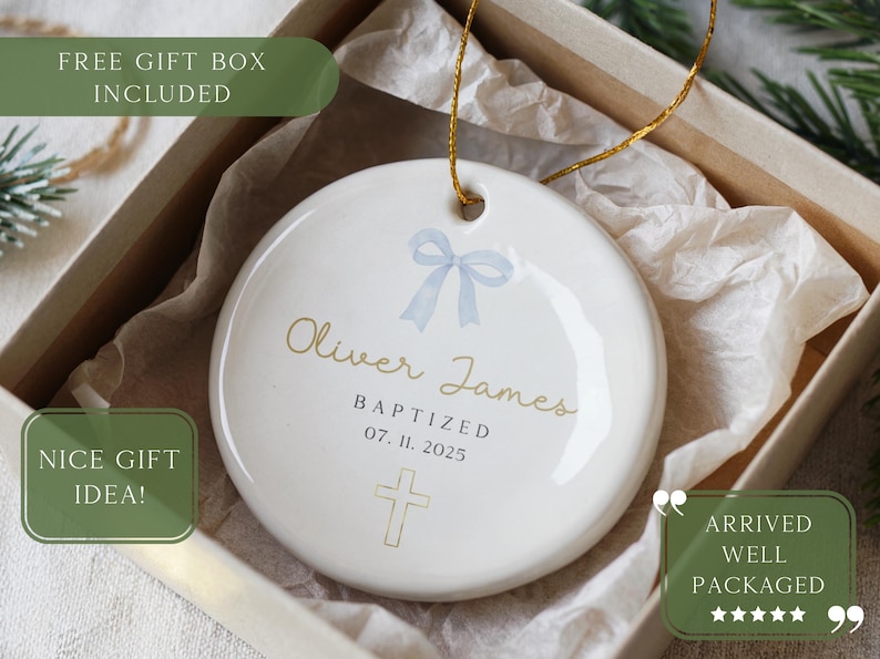 May include: A white ceramic ornament with a gold string, featuring the name "Oliver James" and the date "07. 11. 2025". It includes a light blue bow and a cross design. The ornament is packaged in a gift box.
