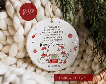 May include: A white ceramic Christmas ornament with a gold string, featuring a festive design. The ornament has text that reads "A job made us coworkers but our sarcasm and inappropriate conversations made us friends. Merry Christmas." Two cartoon geese are wearing Santa hats. The ornament is surrounded by Christmas decorations.