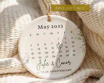 May include: A white ceramic ornament with a calendar design for May 2025, featuring a highlighted date and the names "Julie & Conor" with "10 YEAR ANNIVERSARY" text. The ornament has a gold string for hanging and is decorated with green floral accents.