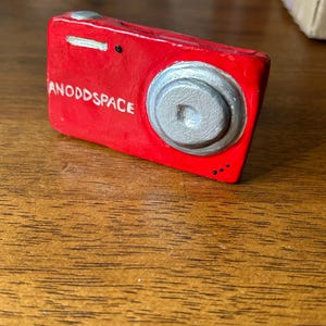 May include: A red toy camera with a silver lens and the words "ANODDSPACE" printed on the front.