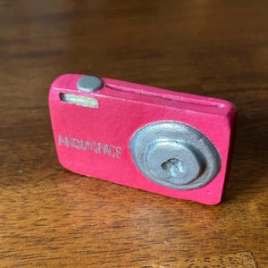 May include: A pink toy camera with a silver lens and the words "ANDDOSPACE" printed on the front.