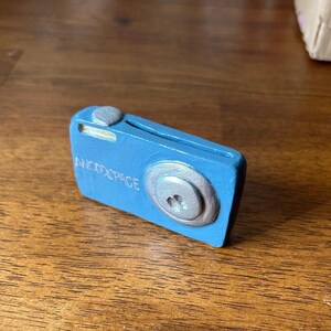 May include: A small, blue camera-shaped object with a silver lens and top button. The text "ANODOSPACE" is printed on the front. The object is likely a decorative item or a small toy.