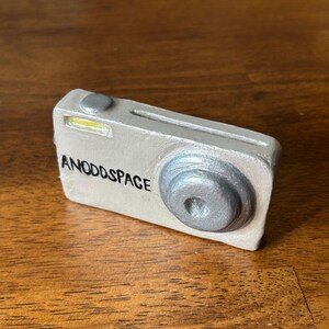 May include: A silver and gray camera with the words "ANODDSPACE" written on the front.