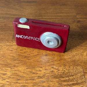 May include: A red camera with a silver lens and a silver button on top. The camera has the words "ANODDSPACE" written on the front.