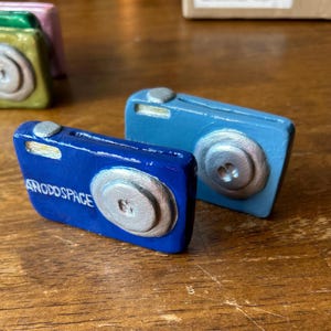 May include: Two miniature toy cameras, one blue and one dark blue, with silver details. The dark blue camera has the text "ANODSPACE" printed on it.