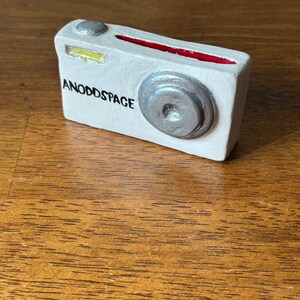 May include: A white camera with a silver lens and a red stripe. The camera has the words "ANODDSPACE" printed on the side.