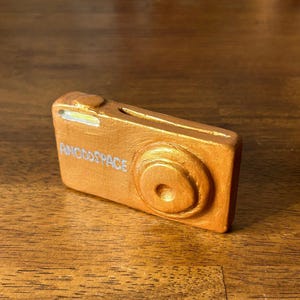 May include: A gold-colored camera shaped object with the text "ANODSPACE" on the front.