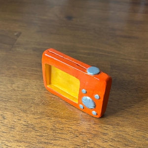 May include: A small, orange, handheld device with a yellow screen and silver buttons. The device is made of clay and has a textured surface.