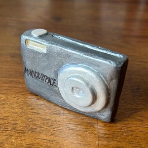 May include: A silver-colored camera with the word "ANODOSPACE" written on the front. The camera has a black lens and a silver button on top.