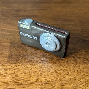 May include: A small, rectangular, handmade camera sculpture with the text "ANODDSPACE" in white. The camera is primarily dark gray with silver and red accents, featuring a lens and a button.