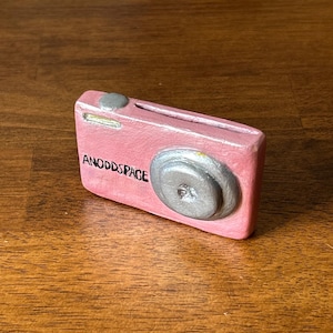 May include: A pink toy camera with a silver lens and the words "ANODDSPACE" written on the front.