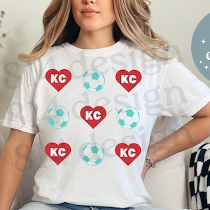 Puede incluir: Una camiseta blanca con un diseño gráfico de corazones rojos y balones de fútbol azules con las letras "KC" dentro de cada corazón. La camiseta la lleva puesta una persona sentada en un sofá blanco.