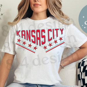 Puede incluir: Camiseta blanca con un gráfico rojo y azul que dice "Kansas City" con estrellas alrededor del texto.
