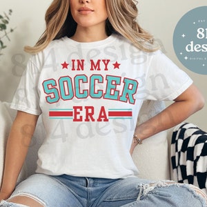 Puede incluir: Camiseta blanca con un gráfico azul y rojo que dice "In My Soccer Era". La camiseta la lleva puesta una persona con el pelo largo rubio y unos vaqueros azules.
