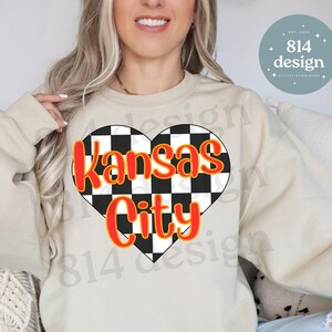 May include: A beige sweatshirt with a black and white checkered heart design that says "Kansas City" in red text with a yellow outline.
