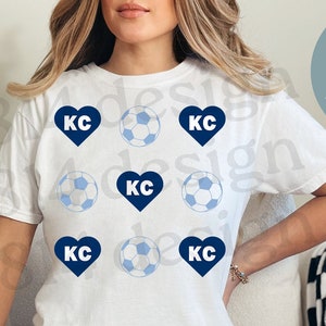 Puede incluir: Una camiseta blanca con un diseño de corazón azul y balón de fútbol. El texto "KC" está dentro del corazón y del balón de fútbol.