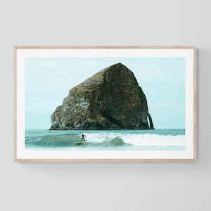 Puede incluir: Una impresión enmarcada que muestra a un surfista en una ola frente a una gran isla rocosa. El mar es turquesa y el cielo está nublado. La impresión está en un marco de madera clara.