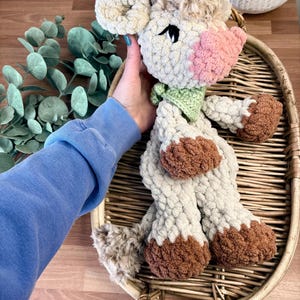 May include: A handmade crochet cow plushie with a light beige body, brown hooves, and a pink snout. The cow has a green scarf and is placed on a woven tray. The plushie has a fluffy head and ears.