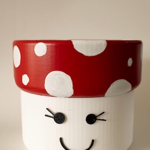 May include: A whimsical, hand-painted mushroom-shaped planter. The top is red with white polka dots, and the base is white with a smiling face and black eyelashes. A charming decorative item for home decor.
