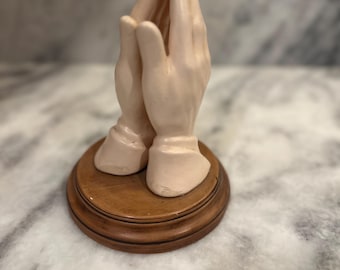 May include: A pair of ceramic hands clasped in prayer, with a wooden base. The hands are a light beige colour.