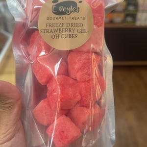 May include: A clear plastic bag filled with freeze-dried strawberry gel cubes. The bag has a label that reads "E. Doyle's Gourmet Treats" and "Freeze Dried Strawberry Gel- OH CUBES". The cubes are bright red and appear to be a snack.