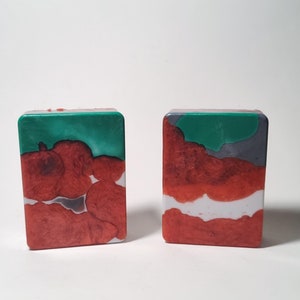 May include: Two square-shaped bars of soap with a marbled design. The soap is red, white, and green.