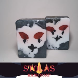 May include: Two bars of black and white hand soap with a red and white design. The soap is shaped like a face with eyes and a nose. The soap is labeled "Stolas Hand Soap".