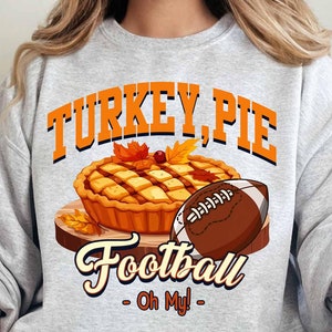 Op de afbeelding: Een lichtgrijs sweatshirt met een grafisch ontwerp. Het ontwerp bevat de woorden "TURKEY, PIE Football - Oh My!" boven een illustratie van een taart en een voetbal. De taart staat op een houten plank met herfstbladeren.