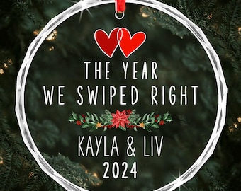 Può includere: Un ornamento natalizio rotondo e trasparente con un nastro rosso. L'ornamento presenta la scritta "THE YEAR WE SWIPED RIGHT" con due cuori rossi, i nomi "KAYLA & LIV" e l'anno "2024". Un design floreale festivo è sottostante.