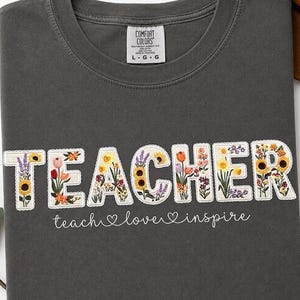 May include: Dark grey t-shirt featuring the word "TEACHER" in white, floral-filled letters. The phrase "teach love inspire" is written below in a handwritten font. The shirt is folded.