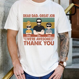 May include: A white t-shirt with a vintage-style graphic featuring a dad and three children. The text reads "Dear Dad, Great Job We're Awesome Thank You". The children's names are Olivia, John, and Liam.