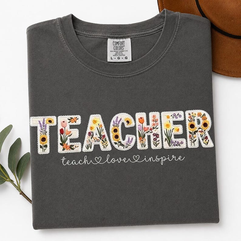 May include: Dark grey t-shirt featuring the word "TEACHER" in white, floral-filled letters. The phrase "teach love inspire" is written below in a handwritten font. The shirt is folded.