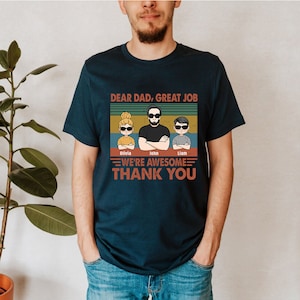 May include: A navy blue t-shirt with a vintage-style graphic that says "Dear Dad, Great Job" with a cartoon image of a dad and two children. The children's names are Olivia, John, and Liam. The graphic also says "We're Awesome" and "Thank You".