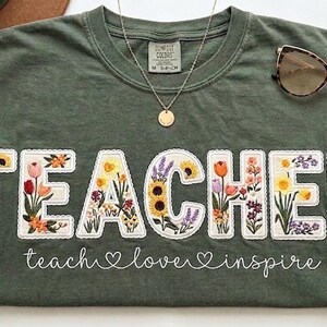 May include: Green t-shirt featuring the word "TEACHER" in white, with each letter filled with floral embroidery. The phrase "teach love inspire" is written below. Sunglasses and a necklace are placed on the shirt.