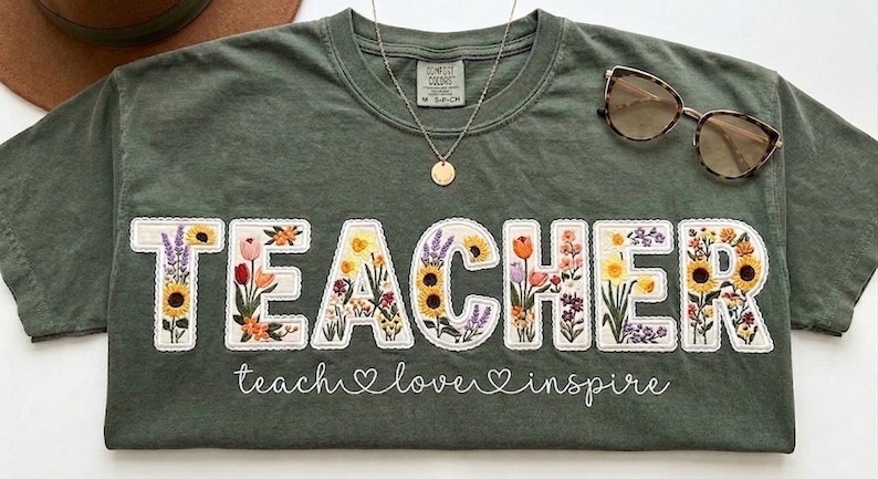 May include: Green t-shirt featuring the word "TEACHER" in white, with each letter filled with floral embroidery. The phrase "teach love inspire" is written below. Sunglasses and a necklace are placed on the shirt.