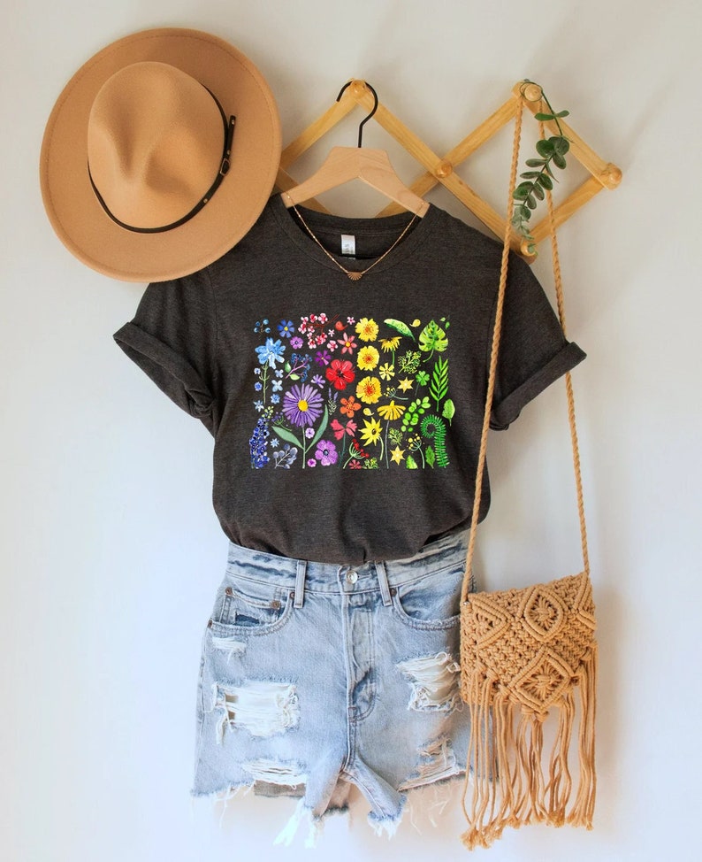 May include: A dark gray t-shirt with a rainbow floral print. The shirt is hanging on a wooden hanger and has a V-neck. The shirt is paired with light blue denim shorts and a brown woven bag.